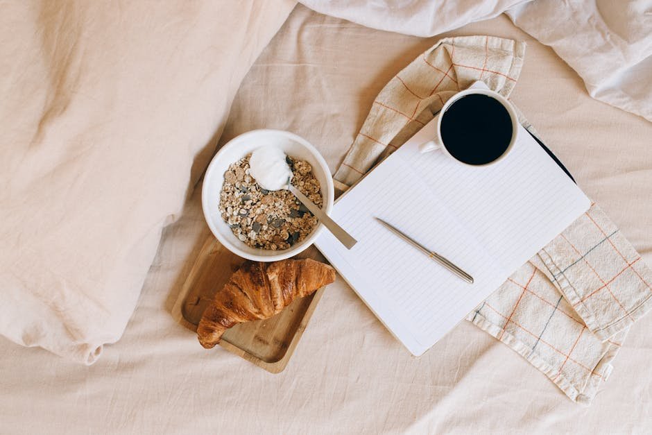 A cozy breakfast setup on a bed featuring a croissant, yogurt bowl, coffee, and notebook.