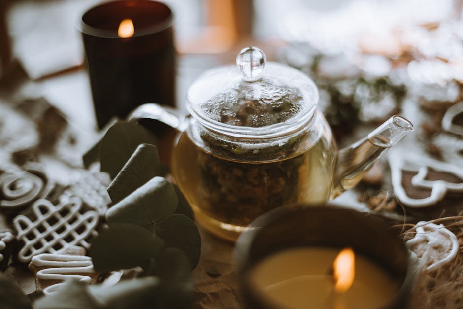 A glass teapot surrounded by candles and green leaves on a rustic wooden table.