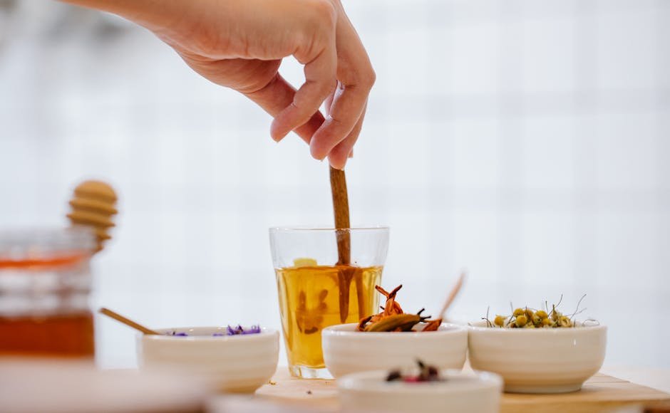 A hand stirs herbal tea in a glass with bowls of spices and herbs on a kitchen counter.